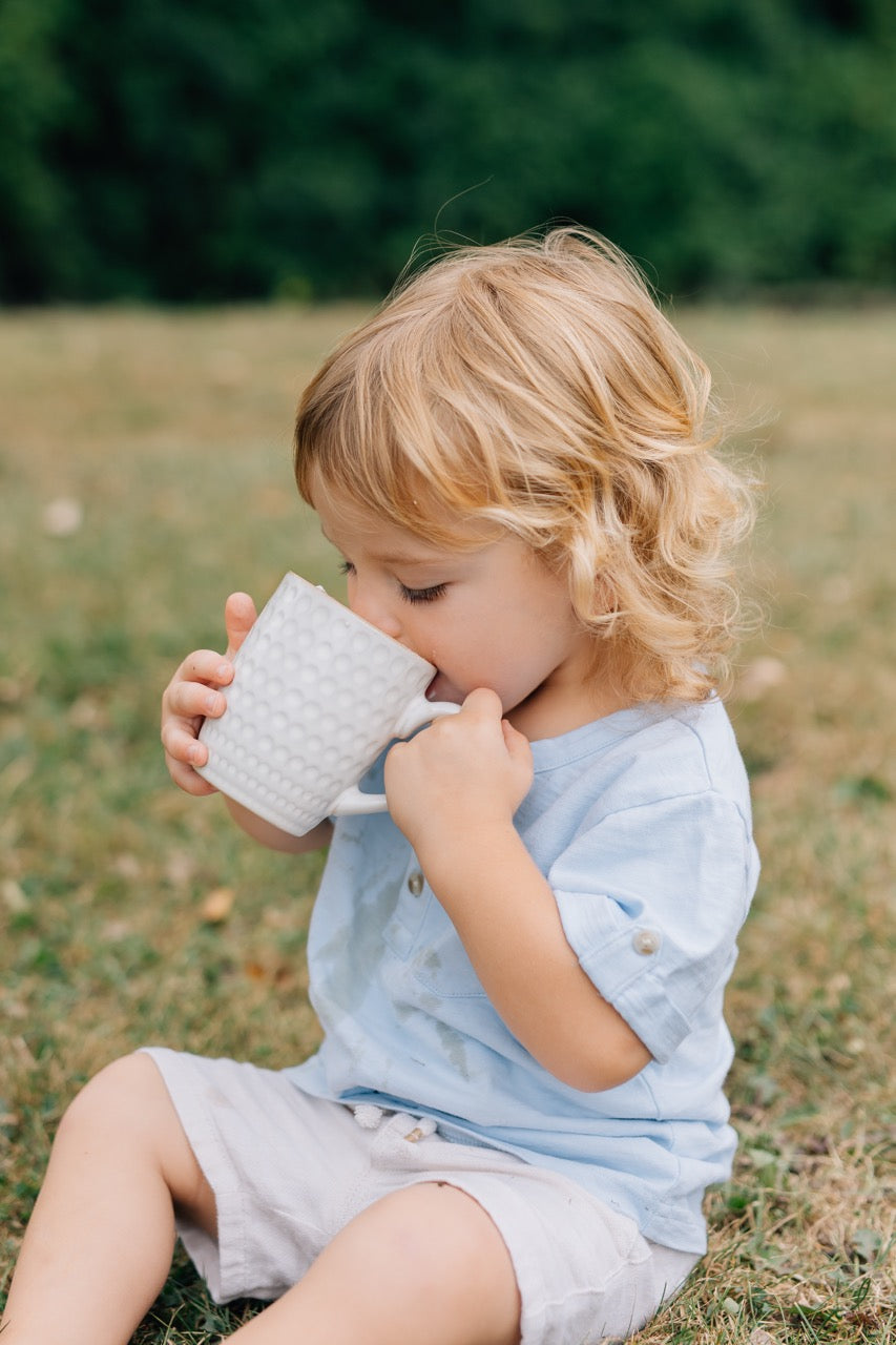 A toddler sits in the grass drinking from a white mug